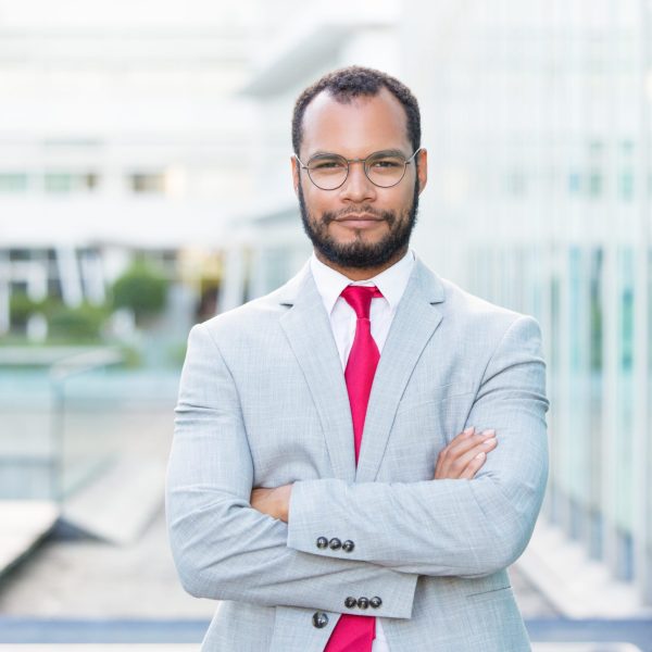 Confident businessman posing outside with folded arms. Handsome young Latin business man wearing eyeglasses and red tie and looking at camera. Successful businessman concept