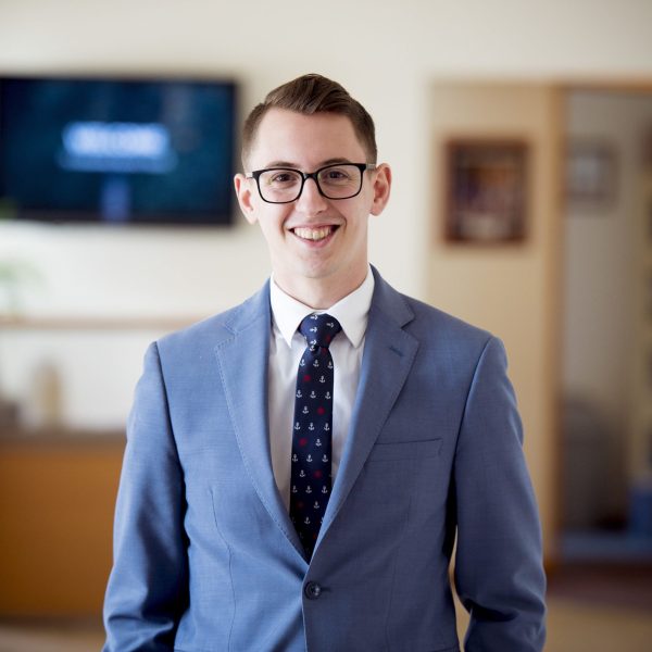 A portrait of a man with glasses in a blue suit with a tie under the lights with a blurry background